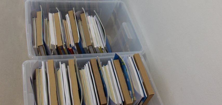 Schulbuchausleihe Lahnstein Mehrere transparente Kunststoffboxen mit nach Klassen sortierten Schulbuchpaketen stehen in einer langen Reihe entlang einer weißen Wand in der Stadtbücherei Lahnstein.