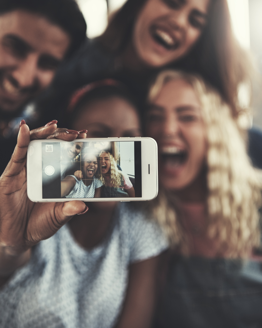 Vier Jugendliche stehen eng beieinander und lachen fröhlich, während sie ein Selfie machen. Im Vordergrund hält eine Person ein Smartphone, auf dessen Display das aufgenommene Gruppenfoto zu sehen ist. Die Stimmung wirkt ausgelassen, freundschaftlich und spontan.