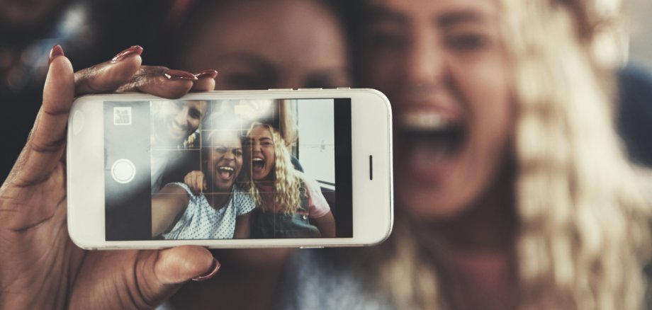 Vier Jugendliche stehen eng beieinander und lachen fröhlich, während sie ein Selfie machen. Im Vordergrund hält eine Person ein Smartphone, auf dessen Display das aufgenommene Gruppenfoto zu sehen ist. Die Stimmung wirkt ausgelassen, freundschaftlich und spontan.