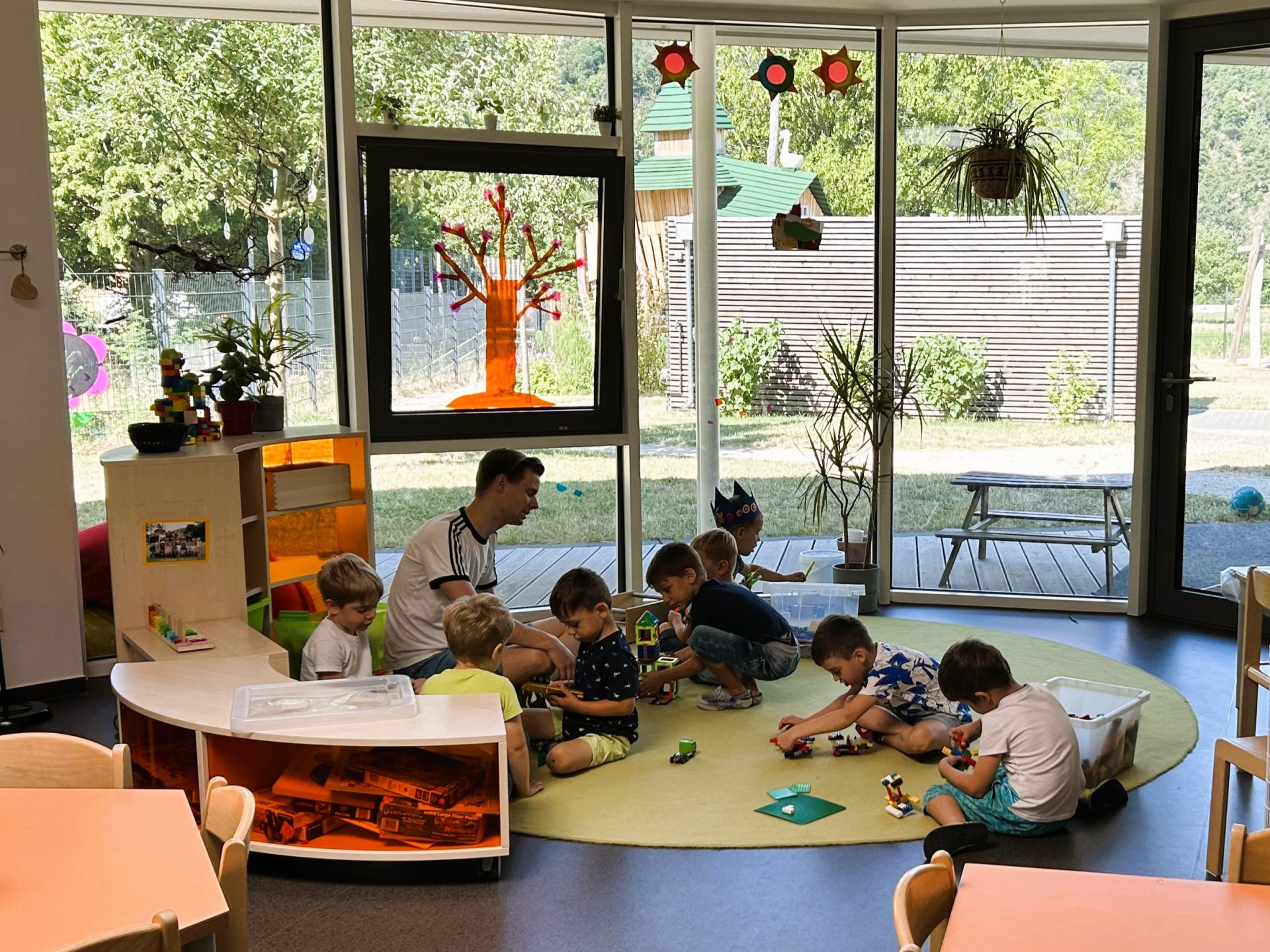 Heller, sonnendurchfluteter Gruppenraum der kommunalen Kita LahnEggs. Ein männlicher Erzieher sitzt mit sechs kleinen Kindern auf einem runden, grünen Teppich und spielt mit Bauklötzen und Spielzeugautos. Der Raum hat große Glasfronten mit Blick auf das Außengelände und einen hölzerne Sichtschutzwand im Hintergrund. An den Fenstern sind bunte, selbstgebastelte Dekorationen wie Sonnen und ein Baum angebracht. Im Vordergrund sind Tische und Regale mit Spielzeug zu sehen.