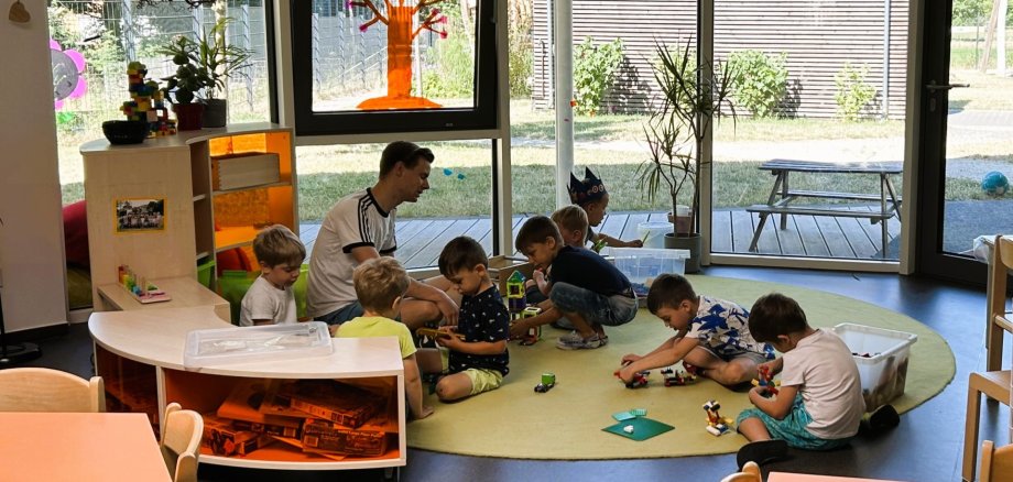 Gruppenraum in der kommunalen Kindertagesstätte LahnEggs Heller, sonnendurchfluteter Gruppenraum der kommunalen Kita LahnEggs. Ein männlicher Erzieher sitzt mit sechs kleinen Kindern auf einem runden, grünen Teppich und spielt mit Bauklötzen und Spielzeugautos. Der Raum hat große Glasfronten mit Blick auf das Außengelände und einen hölzerne Sichtschutzwand im Hintergrund. An den Fenstern sind bunte, selbstgebastelte Dekorationen wie Sonnen und ein Baum angebracht. Im Vordergrund sind Tische und Regale mit Spielzeug zu sehen.