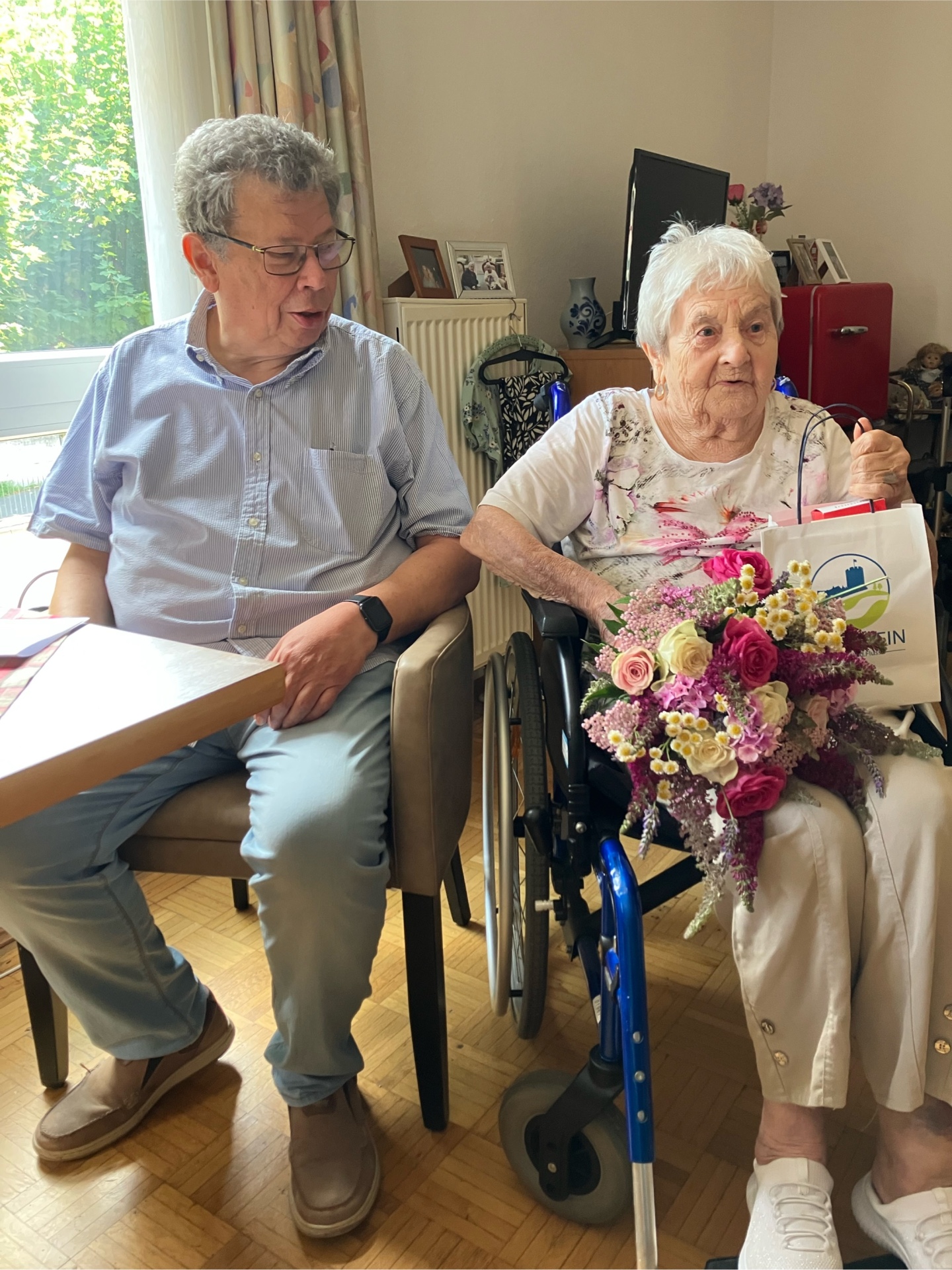 Eine ältere Frau sitzt in einem Rollstuhl in einem gemütlich eingerichteten Wohnraum und hält einen bunten Blumenstrauß sowie eine Geschenktüte in der Hand. Neben ihr sitzt ein Mann auf einem Stuhl und wendet sich ihr zu. Im Hintergrund sind ein Fenster, persönliche Fotos und Möbel zu sehen. Die Atmosphäre wirkt freundlich und feierlich.