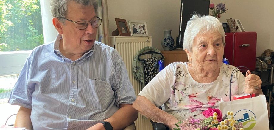 Irmtraudt Fellgiebel feierte ihren 90. Geburtstag in Lahnstein Eine ältere Frau sitzt in einem Rollstuhl in einem gemütlich eingerichteten Wohnraum und hält einen bunten Blumenstrauß sowie eine Geschenktüte in der Hand. Neben ihr sitzt ein Mann auf einem Stuhl und wendet sich ihr zu. Im Hintergrund sind ein Fenster, persönliche Fotos und Möbel zu sehen. Die Atmosphäre wirkt freundlich und feierlich.