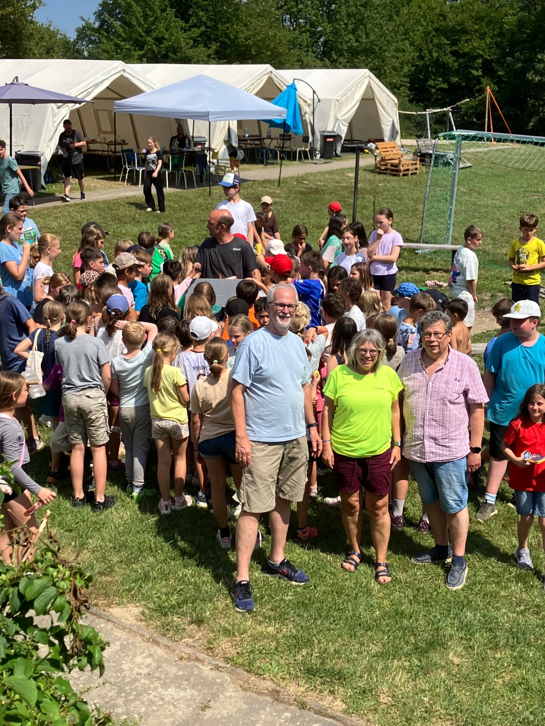 Eine große Gruppe von Kindern versammelt sich auf dem Gelände der AWO-Stadtranderholung. Im Vordergrund stehen Beigeordneter Manfred Radermacher sowie die Leiter Heike und Dirk von Graevenitz. Im Hintergrund sind mehrere weiße Gruppenzelte und spielende Kinder zu sehen.