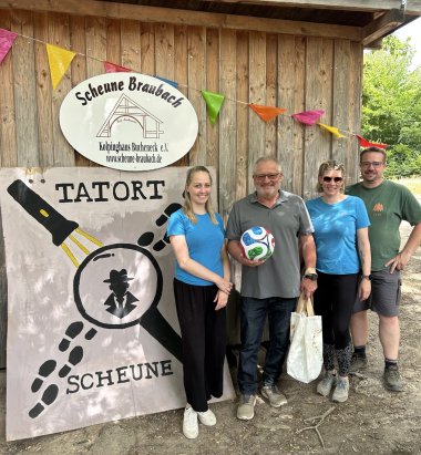 Abwechslungsreiche Ferienangebote für Lahnsteiner Kinder Vier Erwachsene stehen vor einem Holzgebäude mit einem Schild der Ferienfreizeit „Die Scheune“. Bürgermeister Johannes Lauer hält einen Fußball und eine Stofftasche in den Händen. Neben ihm stehen die Leiterinnen und Leiter der Lahnsteiner Gruppe. Im Vordergrund ist ein großes Plakat mit dem Motto „Tatort Scheune“ zu sehen.