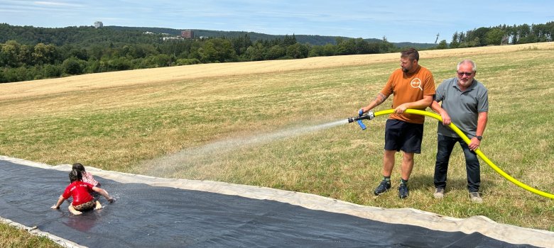 Abwechslungsreiche Ferienangebote für Lahnsteiner Kinder Auf einer großen Wasserrutsche im Freien rutschen zwei Kinder durch das Wasser. Rechts daneben halten OB Lennart Siefert und Bürgermeister Johannes Lauer gemeinsam einen Feuerwehrschlauch und sorgen für ausreichend Wasser. Im Hintergrund sind Wiesen und bewaldete Hügel zu sehen.
