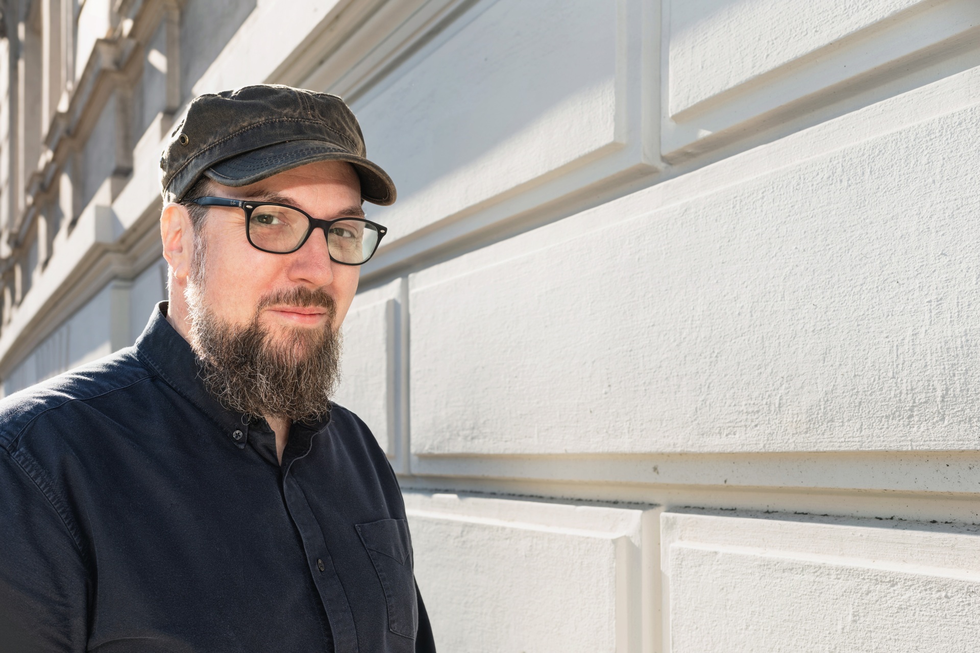 Ein Mann, der Autor Carsten Sebastian Henn, steht vor einer hellen Gebäudefassade und blickt in die Kamera. Er trägt eine dunkle Schirmmütze, eine Brille sowie ein dunkelblaues Hemd. Das Porträt zeigt den Autor in natürlichem Tageslicht.