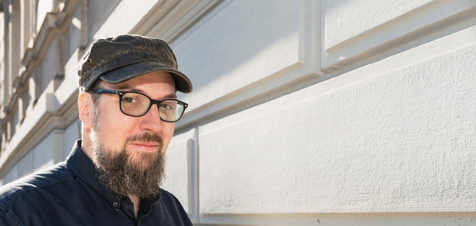 Ein Mann, der Autor Carsten Sebastian Henn, steht vor einer hellen Gebäudefassade und blickt in die Kamera. Er trägt eine dunkle Schirmmütze, eine Brille sowie ein dunkelblaues Hemd. Das Porträt zeigt den Autor in natürlichem Tageslicht.