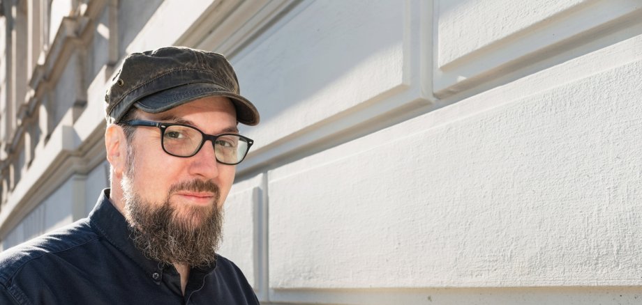 Carsten Sebastian Henn Ein Mann, der Autor Carsten Sebastian Henn, steht vor einer hellen Gebäudefassade und blickt in die Kamera. Er trägt eine dunkle Schirmmütze, eine Brille sowie ein dunkelblaues Hemd. Das Porträt zeigt den Autor in natürlichem Tageslicht.
