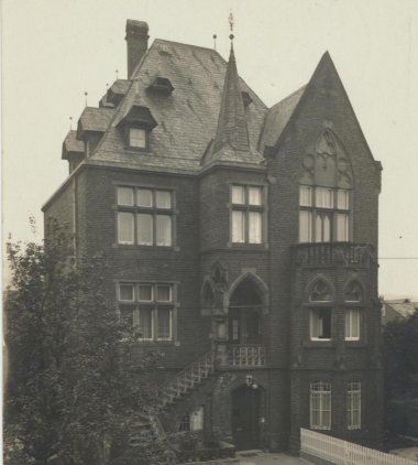 Erkennen Sie Lahnstein? Historische Schwarz-Weiß-Aufnahme eines dreigeschossigen Backsteingebäudes mit steilem Dach und neugotischen Gestaltungselementen. Eine Freitreppe führt zum erhöhten Eingangsportal, vor dem Haus befinden sich ein gepflasterter Hof, Bäume und ein niedriger Zaun.