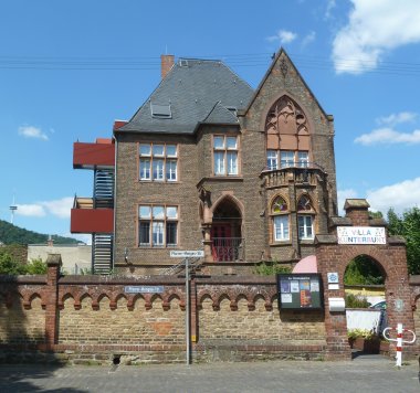 Erkennen Sie Lahnstein? Farbaufnahme eines dreigeschossigen Backsteingebäudes mit steilem Schieferdach, großen Fenstern, Spitzbogenportal und Balkon. Links schließt ein moderner Treppenanbau an das Gebäude an. Vor dem Grundstück stehen eine Backsteinmauer mit Tor, ein Hinweisschild „Villa Kunterbunt“ und ein Schaukasten. Im Vordergrund verläuft eine Straße, darüber spannt sich ein blauer Himmel mit einzelnen Wolken.