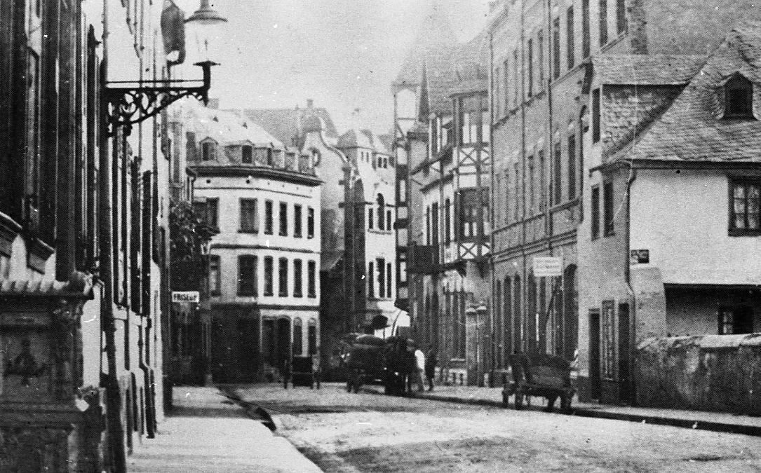 Historische Schwarz-Weiß-Aufnahme einer schmalen Straße mit mehrgeschossigen Wohn- und Geschäftshäusern auf beiden Seiten. Die Gebäude weisen unterschiedliche Fassadenformen auf, darunter verputzte Häuser und Fachwerk. Im Vordergrund hängt eine Straßenlaterne an der Hauswand. Auf der Straße stehen ein Pferdewagen und ein Handkarren, vereinzelt sind Personen zu erkennen. Die Straße wirkt ruhig und vermittelt den Eindruck eines historischen Stadtbildes.