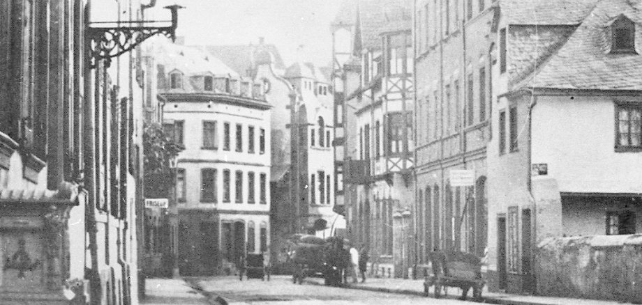 Erkennen Sie Lahnstein? Historische Schwarz-Weiß-Aufnahme einer schmalen Straße mit mehrgeschossigen Wohn- und Geschäftshäusern auf beiden Seiten. Die Gebäude weisen unterschiedliche Fassadenformen auf, darunter verputzte Häuser und Fachwerk. Im Vordergrund hängt eine Straßenlaterne an der Hauswand. Auf der Straße stehen ein Pferdewagen und ein Handkarren, vereinzelt sind Personen zu erkennen. Die Straße wirkt ruhig und vermittelt den Eindruck eines historischen Stadtbildes.