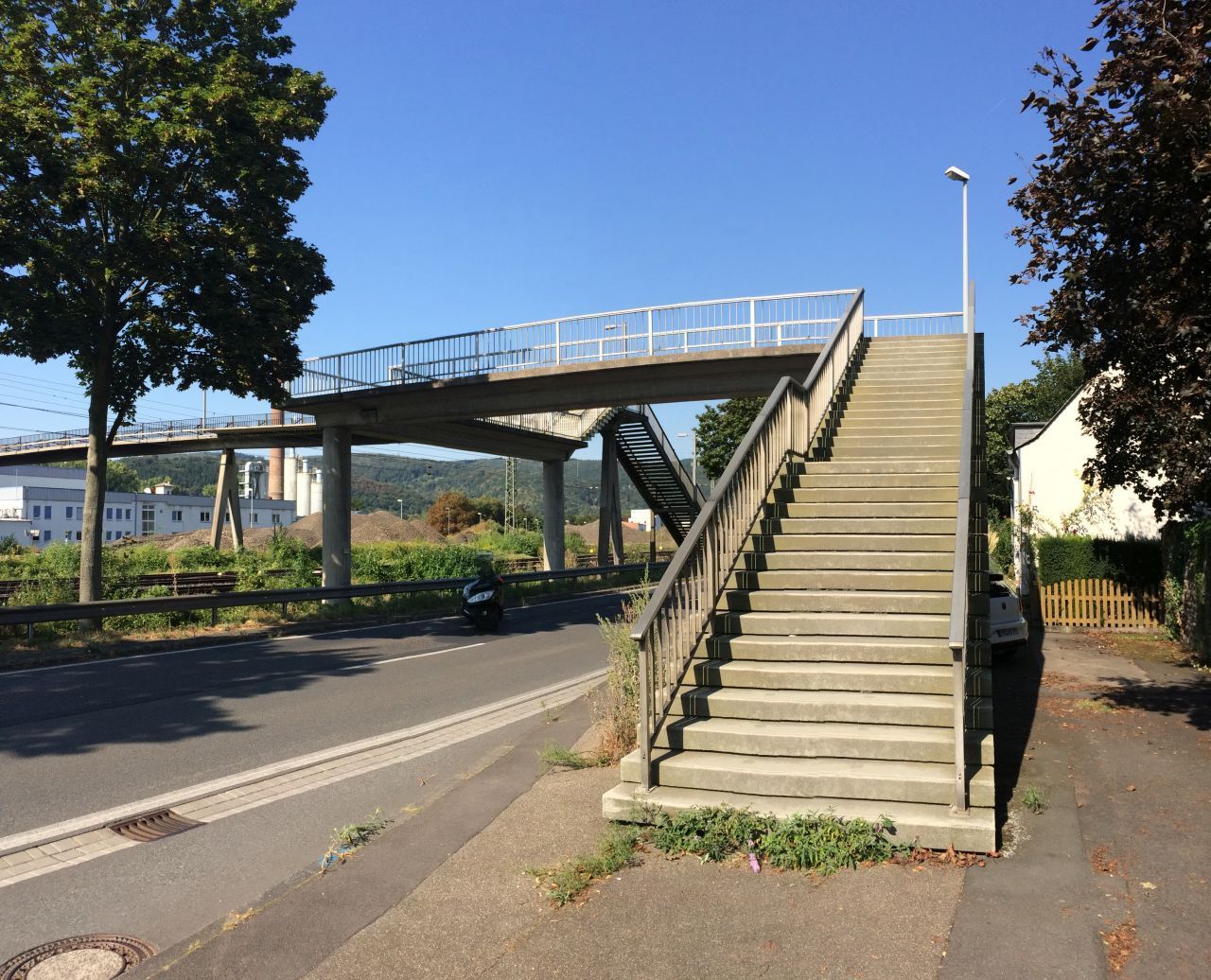 Eine Fußgängerbrücke aus Beton und Stahl überspannt eine Straße sowie die Bahngleise in Oberlahnstein. Im Vordergrund führt eine breite Treppe mit Geländer zur Brücke hinauf. Die Aufnahme entstand bei sonnigem Wetter mit blauem Himmel; links und rechts säumen Bäume den Straßenrand.