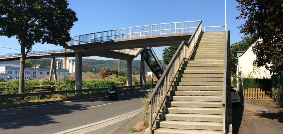 Eine Fußgängerbrücke aus Beton und Stahl überspannt eine Straße sowie die Bahngleise in Oberlahnstein. Im Vordergrund führt eine breite Treppe mit Geländer zur Brücke hinauf. Die Aufnahme entstand bei sonnigem Wetter mit blauem Himmel; links und rechts säumen Bäume den Straßenrand.