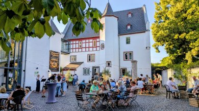 Das Foto zeigt den Innenhof vor dem Theater Lahnstein an einem sonnigen Sommerabend. Im Mittelpunkt steht der historische Sporkenburger Hof mit seiner weiß verputzten Fassade, roten Fachwerkelementen, mehreren kleinen Fenstern und markanten Rundtürmen mit spitzen Schieferdächern. Links schließt sich der moderne Glasbau des Theaters an. Über dem Hof sind Lichterketten gespannt, die zur gemütlichen Atmosphäre beitragen. Im Vordergrund und auf dem gesamten Platz sitzen zahlreiche Besucherinnen und Besucher an Holztischen und schwarzen Stühlen oder stehen in kleinen Gruppen zusammen und unterhalten sich. Einige genießen Getränke oder Speisen, andere verfolgen eine Sängerin, die links auf einer kleinen Bühne mit Mikrofon auftritt. Vor der Bühne steht ein Stehtisch, an dem sich Gäste versammelt haben. Große Laubbäume rahmen den Hof ein und spenden Schatten. Das warme Abendlicht taucht Gebäude, Bäume und Menschen in eine freundliche, sommerliche Stimmung und vermittelt den Eindruck eines gut besuchten, entspannten Stadtfestes.