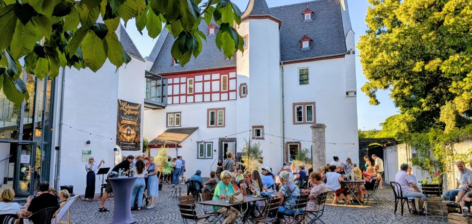 Lahnsteiner Treff lockte rund 500 Gäste vor das Stadttheater Das Foto zeigt den Innenhof vor dem Theater Lahnstein an einem sonnigen Sommerabend. Im Mittelpunkt steht der historische Sporkenburger Hof mit seiner weiß verputzten Fassade, roten Fachwerkelementen, mehreren kleinen Fenstern und markanten Rundtürmen mit spitzen Schieferdächern. Links schließt sich der moderne Glasbau des Theaters an. Über dem Hof sind Lichterketten gespannt, die zur gemütlichen Atmosphäre beitragen. Im Vordergrund und auf dem gesamten Platz sitzen zahlreiche Besucherinnen und Besucher an Holztischen und schwarzen Stühlen oder stehen in kleinen Gruppen zusammen und unterhalten sich. Einige genießen Getränke oder Speisen, andere verfolgen eine Sängerin, die links auf einer kleinen Bühne mit Mikrofon auftritt. Vor der Bühne steht ein Stehtisch, an dem sich Gäste versammelt haben. Große Laubbäume rahmen den Hof ein und spenden Schatten. Das warme Abendlicht taucht Gebäude, Bäume und Menschen in eine freundliche, sommerliche Stimmung und vermittelt den Eindruck eines gut besuchten, entspannten Stadtfestes.