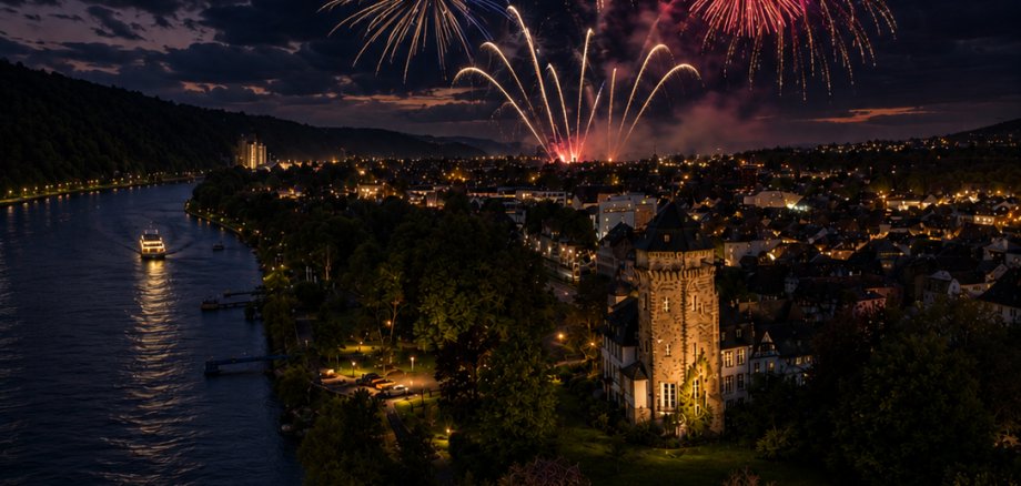 Ein großes Feuerwerk erleuchtet den Nachthimmel über einer Stadt am Rhein. Mehrere farbenfrohe Feuerwerkskörper explodieren in Gold-, Rot- und Weißtönen über den Dächern der beleuchteten Altstadt. Im Vordergrund ist ein historischer Turm von einem Park umgeben, links fließt der Rhein, auf dem ein beleuchtetes Fahrgastschiff unterwegs ist. Dunkle Wolken ziehen über den Himmel und bilden einen eindrucksvollen Kontrast zu den hellen Feuerwerkslichtern.