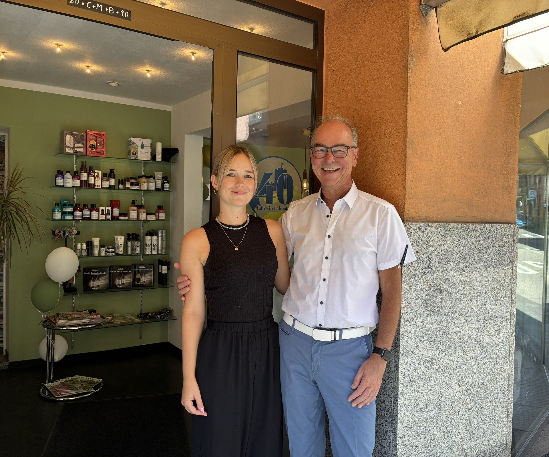 Vor dem Eingang eines Friseursalons stehen zwei Personen nebeneinander und blicken lächelnd in die Kamera. Links steht eine junge Frau mit schulterlangem blondem Haar, einem schwarzen ärmellosen Oberteil und einer schwarzen Hose. Rechts steht ein älterer Mann mit Brille, weißem Kurzarmhemd und hellblauer Hose; er hat den Arm freundschaftlich um die Frau gelegt. Hinter ihnen ist durch die geöffnete Glastür der Salon mit Regalen voller Haarpflegeprodukte zu sehen. Auf der Glastür befindet sich ein rundes Schild mit der Aufschrift „40 Jahre in Lahnstein“, das Lahnstein-Label zum Jubiläum des Betriebs.