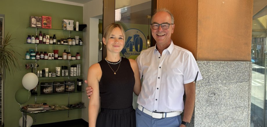 40 Jahre Hairworld Moritz in Lahnstein Vor dem Eingang eines Friseursalons stehen zwei Personen nebeneinander und blicken lächelnd in die Kamera. Links steht eine junge Frau mit schulterlangem blondem Haar, einem schwarzen ärmellosen Oberteil und einer schwarzen Hose. Rechts steht ein älterer Mann mit Brille, weißem Kurzarmhemd und hellblauer Hose; er hat den Arm freundschaftlich um die Frau gelegt. Hinter ihnen ist durch die geöffnete Glastür der Salon mit Regalen voller Haarpflegeprodukte zu sehen. Auf der Glastür befindet sich ein rundes Schild mit der Aufschrift „40 Jahre in Lahnstein“, das Lahnstein-Label zum Jubiläum des Betriebs.