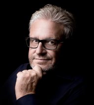 Porträtaufnahme einer Person mit kurzen grauen Haaren, schwarzer Brille und leichtem Bart. Die Person trägt ein dunkles Oberteil, blickt direkt in die Kamera und stützt das Kinn nachdenklich auf die Hand. Der schwarze Hintergrund und die gezielte Ausleuchtung lenken den Fokus auf das Gesicht und verleihen dem Bild eine ruhige, professionelle Wirkung.