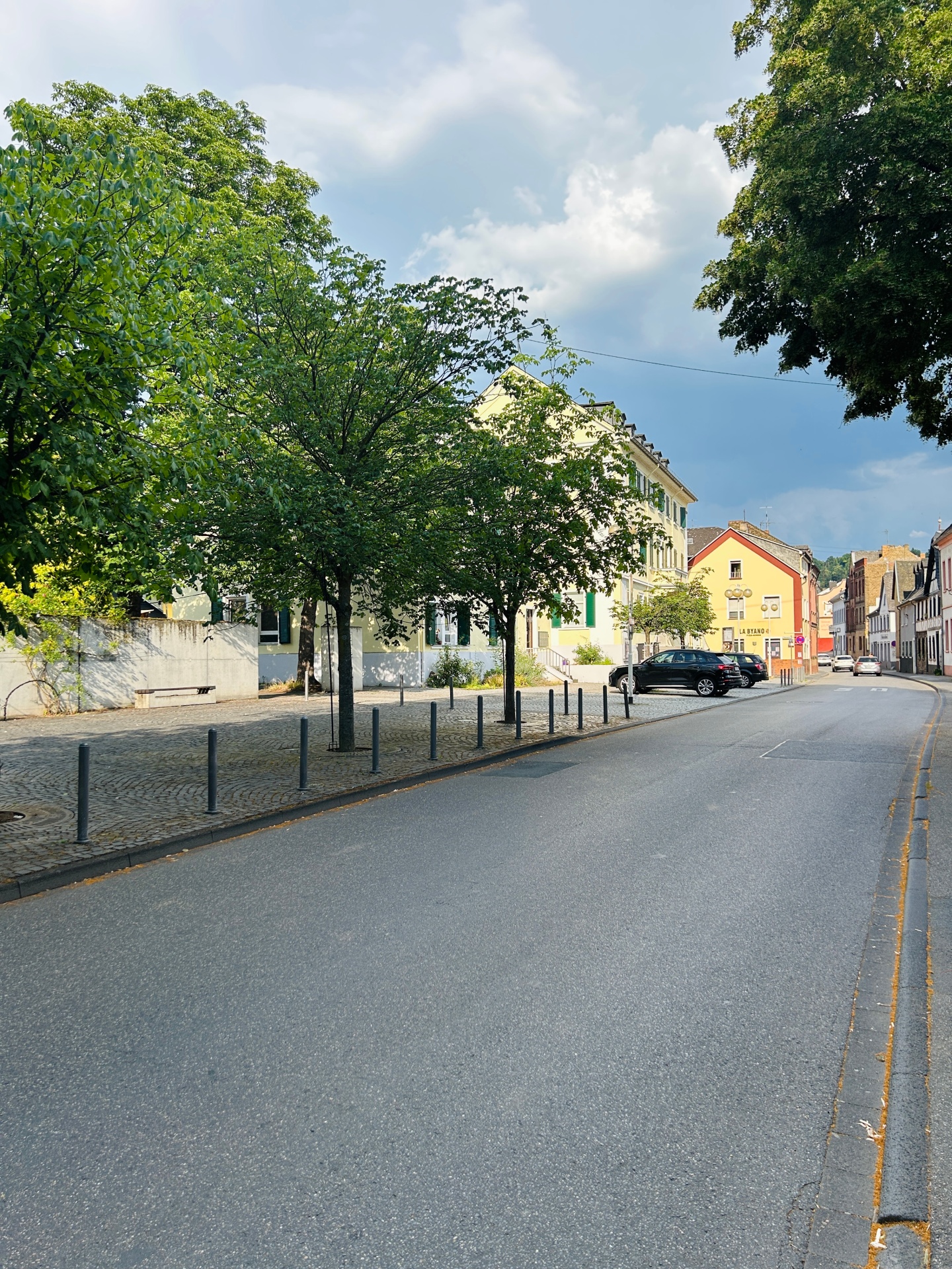 Blick entlang einer ruhigen Straße in Lahnstein. Auf der linken Seite stehen mehrere große, dicht belaubte Bäume vor einem hellen Gebäude und einem gepflasterten Platz mit Pollern. Rechts verläuft die Straße durch eine Reihe historischer Wohn- und Geschäftshäuser. Im Hintergrund ziehen dunkle Wolken auf, während die Szenerie noch von Sonnenlicht erhellt wird.