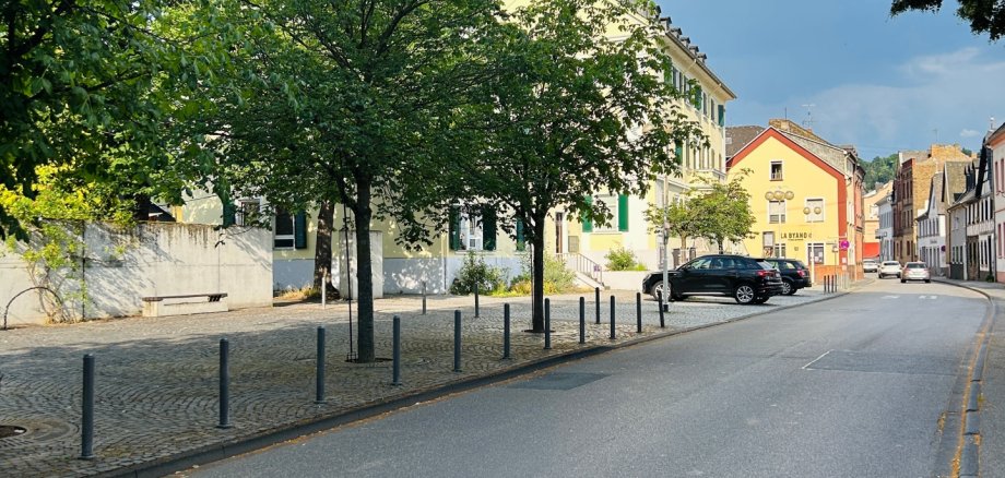 Blick entlang einer ruhigen Straße in Lahnstein. Auf der linken Seite stehen mehrere große, dicht belaubte Bäume vor einem hellen Gebäude und einem gepflasterten Platz mit Pollern. Rechts verläuft die Straße durch eine Reihe historischer Wohn- und Geschäftshäuser. Im Hintergrund ziehen dunkle Wolken auf, während die Szenerie noch von Sonnenlicht erhellt wird.