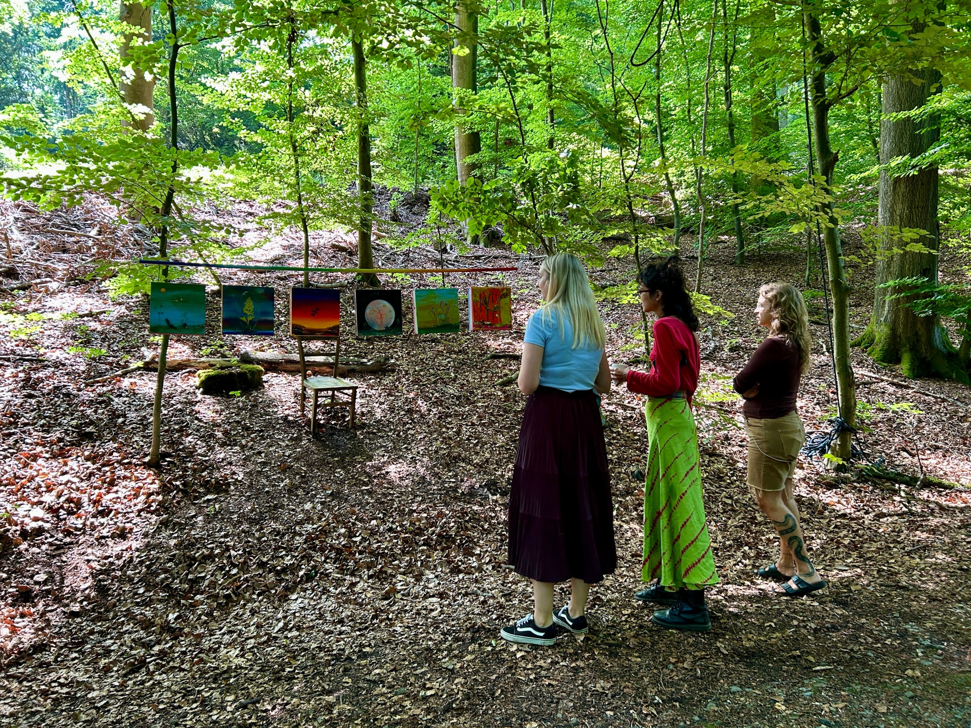Drei Frauen betrachten im Wald eine Outdoor-Kunstausstellung mit mehreren farbenfrohen Bildern, die zwischen Bäumen aufgehängt sind. Sonnenlicht fällt durch das dichte Blätterdach auf den Waldboden und schafft eine ruhige, natürliche Atmosphäre.