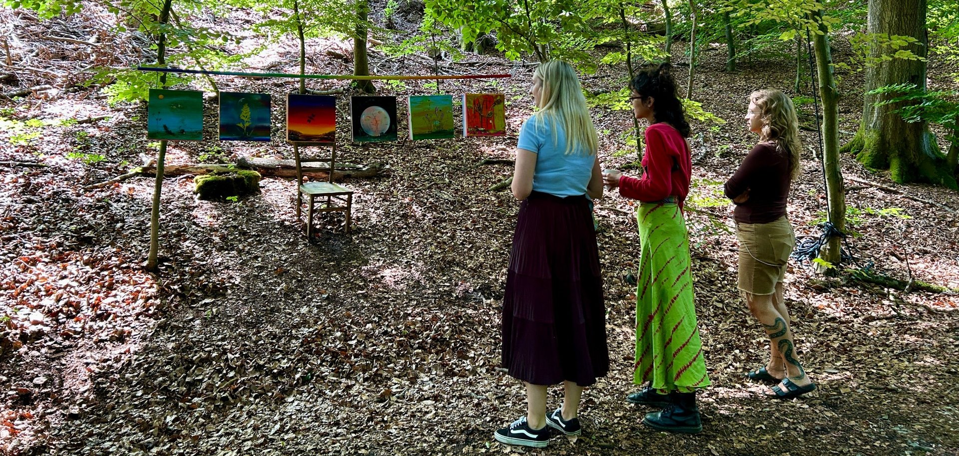 Drei Frauen betrachten im Wald eine Outdoor-Kunstausstellung mit mehreren farbenfrohen Bildern, die zwischen Bäumen aufgehängt sind. Sonnenlicht fällt durch das dichte Blätterdach auf den Waldboden und schafft eine ruhige, natürliche Atmosphäre.