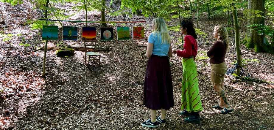 Drei Frauen betrachten im Wald eine Outdoor-Kunstausstellung mit mehreren farbenfrohen Bildern, die zwischen Bäumen aufgehängt sind. Sonnenlicht fällt durch das dichte Blätterdach auf den Waldboden und schafft eine ruhige, natürliche Atmosphäre.
