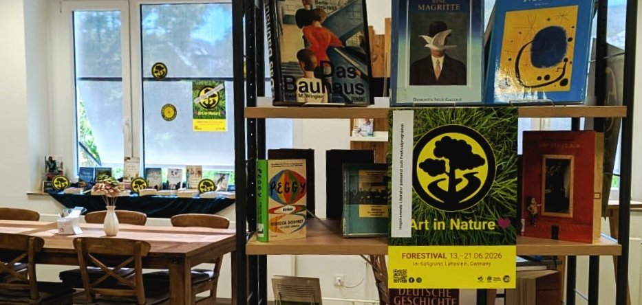 Ein Bücherregal in der Stadtbücherei Lahnstein mit thematisch präsentierten Büchern zu Kunst, Geschichte und den „Goldenen Zwanzigern“. Zwischen den Büchern hängen mehrere gelb-schwarze „Forestival“-Plakate und Aufkleber. Im Hintergrund sind Sitzplätze und große Fenster der Bücherei zu sehen.