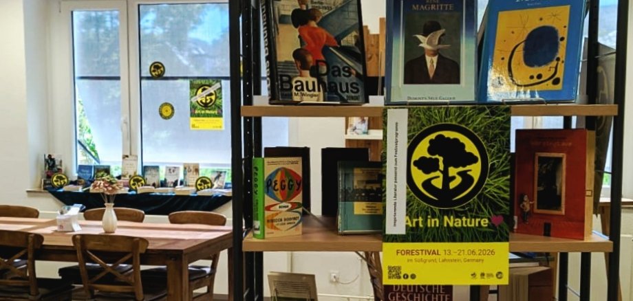 Forestival trifft auf Stadtbücherei Lahnstein Ein Bücherregal in der Stadtbücherei Lahnstein mit thematisch präsentierten Büchern zu Kunst, Geschichte und den „Goldenen Zwanzigern“. Zwischen den Büchern hängen mehrere gelb-schwarze „Forestival“-Plakate und Aufkleber. Im Hintergrund sind Sitzplätze und große Fenster der Bücherei zu sehen.