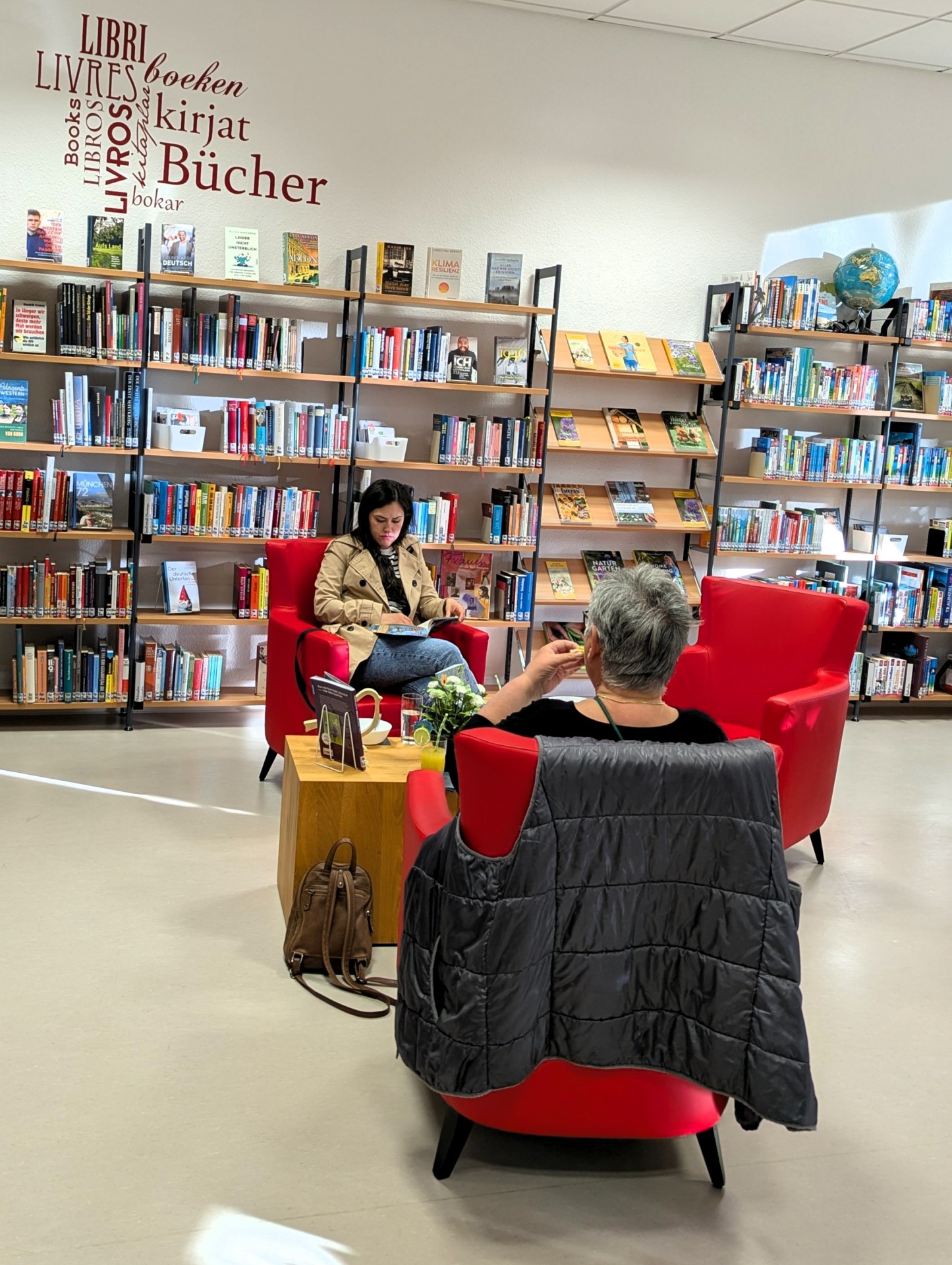 Zwei Frauen sitzen in roten Sesseln der Stadtbücherei Lahnstein und lesen in entspannter Atmosphäre. Umgeben von hohen Bücherregalen, kleinen Tischen mit Getränken und warmem Licht entsteht eine ruhige und gemütliche Stimmung zwischen den Büchern.
