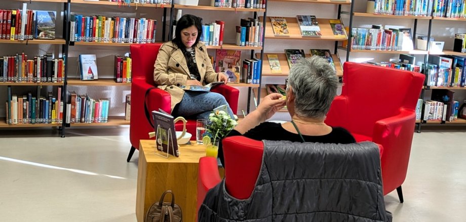 Zwei Frauen sitzen in roten Sesseln der Stadtbücherei Lahnstein und lesen in entspannter Atmosphäre. Umgeben von hohen Bücherregalen, kleinen Tischen mit Getränken und warmem Licht entsteht eine ruhige und gemütliche Stimmung zwischen den Büchern.
