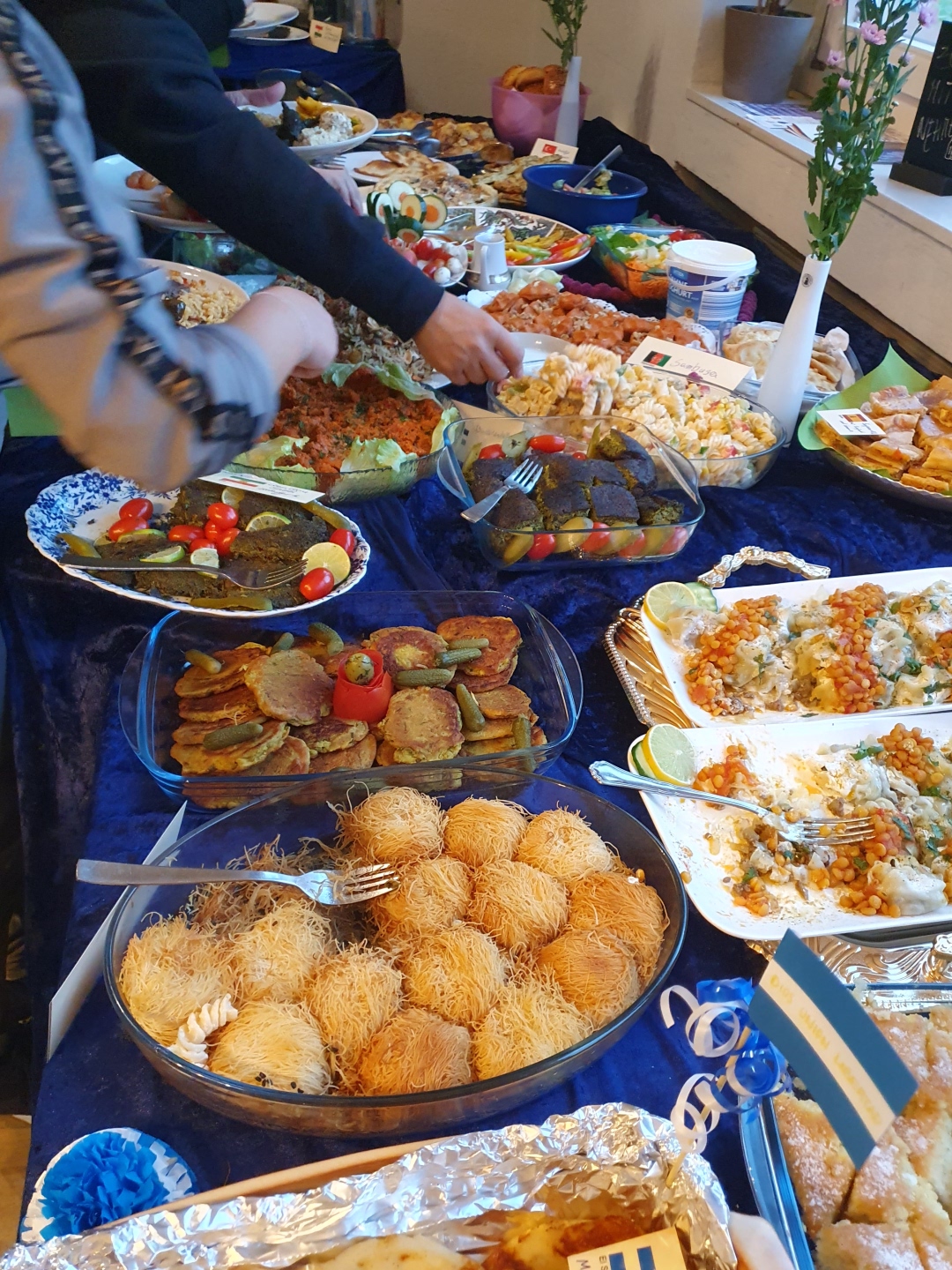 Ein großes internationales Buffet mit zahlreichen herzhaften und süßen Speisen aus verschiedenen Ländern. Mehrere Menschen bedienen sich an den liebevoll angerichteten Gerichten auf einem langen Tisch mit blauer Tischdecke.