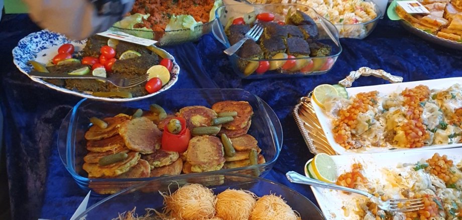 Eine-Welt-Dinner in Lahnstein Ein großes internationales Buffet mit zahlreichen herzhaften und süßen Speisen aus verschiedenen Ländern. Mehrere Menschen bedienen sich an den liebevoll angerichteten Gerichten auf einem langen Tisch mit blauer Tischdecke.