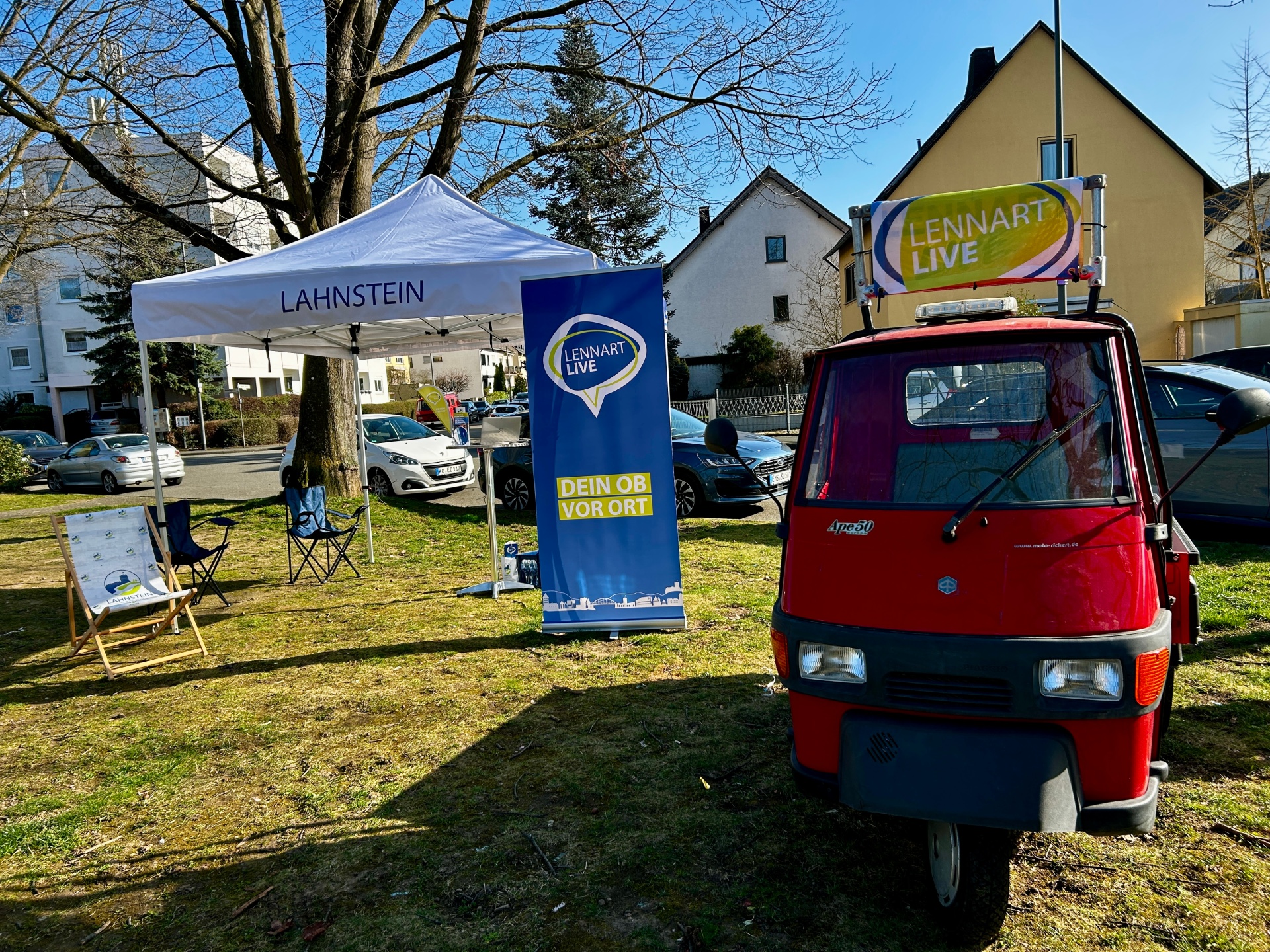 Rote Ape mit „Lennart Live“-Beschilderung steht neben einem weißen Pavillon der Stadt Lahnstein auf einer Grünfläche in einem Wohngebiet. Vor Ort sind Sitzgelegenheiten und ein Banner mit der Aufschrift „Dein OB vor Ort“ aufgebaut.