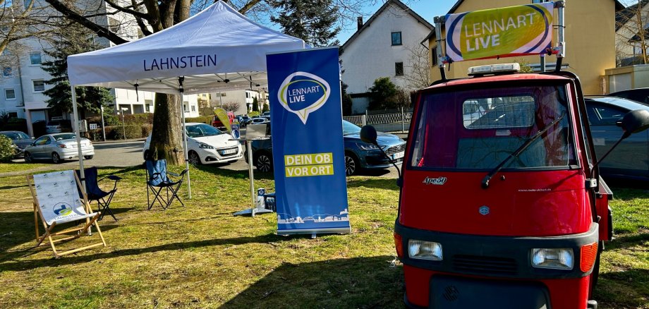 Einwohnersprechstunde Lennart Live Rote Ape mit „Lennart Live“-Beschilderung steht neben einem weißen Pavillon der Stadt Lahnstein auf einer Grünfläche in einem Wohngebiet. Vor Ort sind Sitzgelegenheiten und ein Banner mit der Aufschrift „Dein OB vor Ort“ aufgebaut.