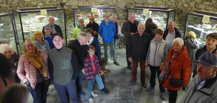 Museumstag Eine große Gruppe von Menschen unterschiedlichen Alters steht in einem historischen Kellergewölbe mit Steinmauern, in dem archäologische Fundstücke in beleuchteten Glasvitrinen ausgestellt sind.
