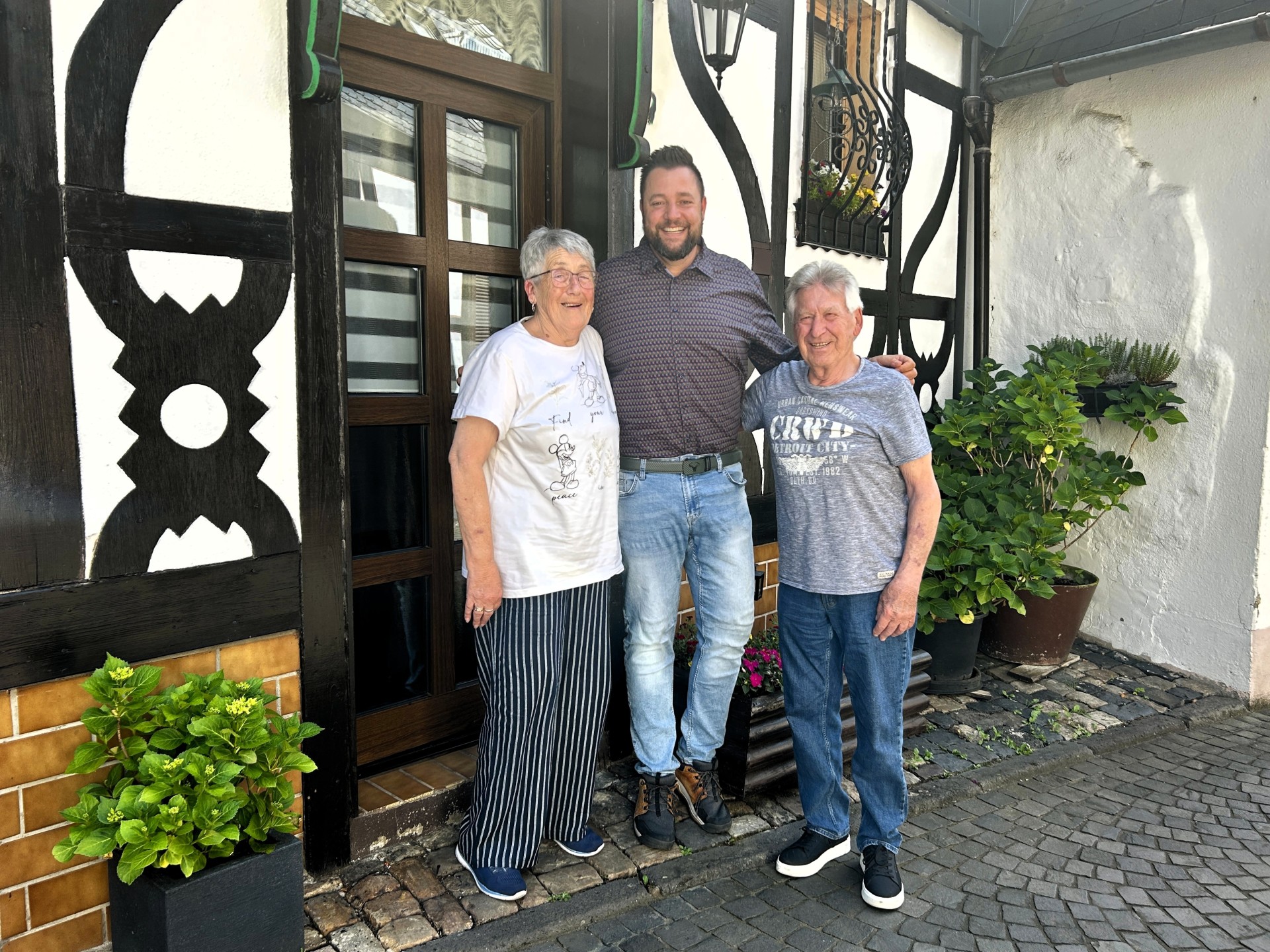 Drei Personen stehen lächelnd vor einem Fachwerkhaus mit dunklen Holzbalken und weißer Fassade. In der Mitte steht ein jüngerer Mann mit einem Arm um die Schultern eines älteren Paares, das links und rechts neben ihm steht. Die Frau trägt ein helles T-Shirt und eine gestreifte Hose, der Mann ein graues T-Shirt und Jeans. Neben der Haustür und auf dem Boden stehen mehrere grüne Pflanzen in Töpfen, und der gepflasterte Boden verstärkt die gemütliche, ländliche Atmosphäre.