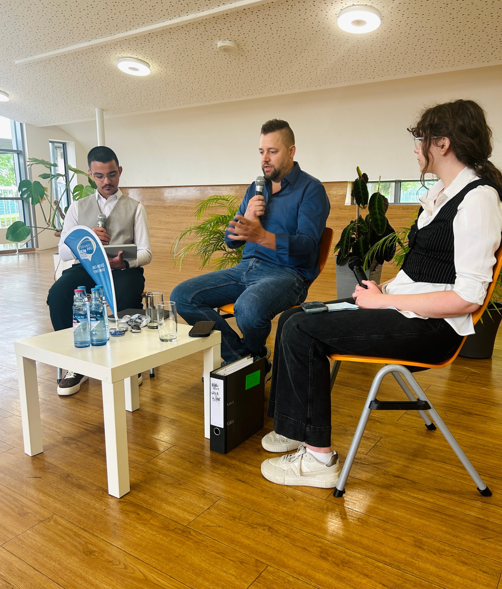 Eine Nahaufnahme zeigt Lennart Siefert (Mitte) in einem blauen Hemd, wie er auf einem Stuhl auf der Bühne sitzt und in ein Mikrofon spricht. Rechts sitzt Janja Stefanac in einer weißen Bluse und schwarzer Weste und hält ebenfalls ein Mikrofon. Links sitzt Abdul Salkho in einem hellen Hemd und beiger Weste, der ein Tablet und ein Mikrofon hält. Im Vordergrund stehen Wasserflaschen, eine Sonnenbrille und ein kleiner Wimpel auf einem Tischchen, sowie ein schwarzer Ordner auf dem Boden.