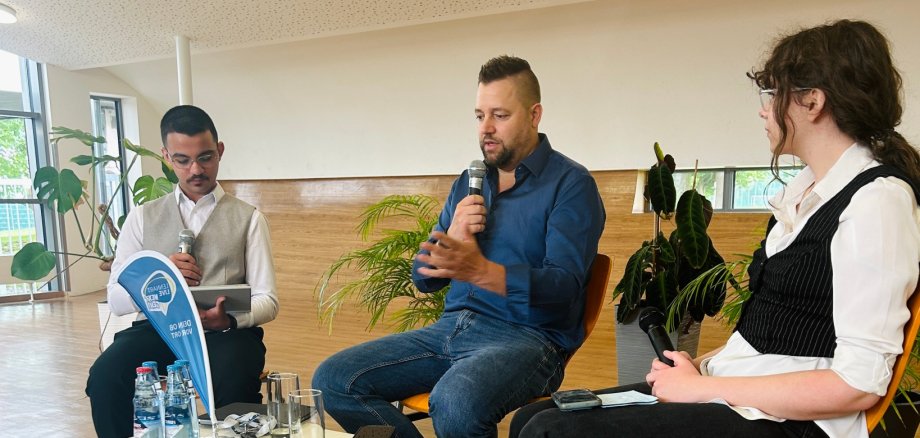 Jugendsprechstunde am MDG Eine Nahaufnahme zeigt Lennart Siefert (Mitte) in einem blauen Hemd, wie er auf einem Stuhl auf der Bühne sitzt und in ein Mikrofon spricht. Rechts sitzt Janja Stefanac in einer weißen Bluse und schwarzer Weste und hält ebenfalls ein Mikrofon. Links sitzt Abdul Salkho in einem hellen Hemd und beiger Weste, der ein Tablet und ein Mikrofon hält. Im Vordergrund stehen Wasserflaschen, eine Sonnenbrille und ein kleiner Wimpel auf einem Tischchen, sowie ein schwarzer Ordner auf dem Boden.