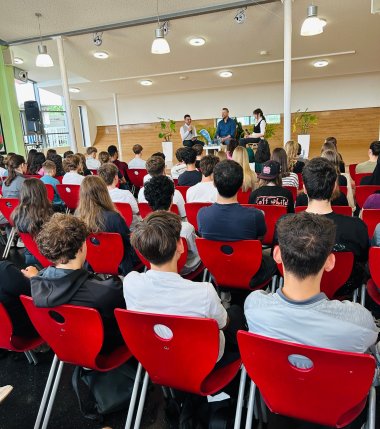 Jugendsprechstunde am MDG Blick von hinten in eine voll besetzte Schulaula. Schülerinnen und Schüler sitzen in langen Reihen auf roten Stühlen und blicken nach vorne auf eine Bühne. Auf der Bühne sind drei Personen zu sehen, darunter Oberbürgermeister Lennart Siefert (Mitte), der in ein Mikrofon spricht, flankiert von Janja Stefanac und Abdul Salkho. Zimmerpflanzen flankieren die Bühne.
