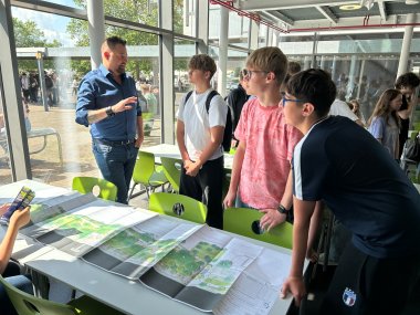 Jugendsprechstunde am MDG Ein helles Foto zeigt Oberbürgermeister Lennart Siefert (links) in einem blauen Hemd und Jeans, wie er mit drei Schülern an einem Tisch in einer Mensa spricht. Er gestikuliert, während die Schüler, darunter ein Junge mit rosa T-Shirt und Brille, aufmerksam zuhören. Auf dem Tisch vor ihnen liegt ein detaillierter Bebauungsplan ausgebreitet.