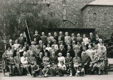 Vom Gesellenverein zur Kolpingfamilie Ein historisches Schwarz-Weiß-Gruppenfoto vor einer Natursteinkirche. Zahlreiche Frauen, Männer und Kinder in festlicher Kleidung aus einer früheren Zeit sind in mehreren Reihen aufgestellt und sitzen teilweise auf Stühlen oder auf dem Boden.