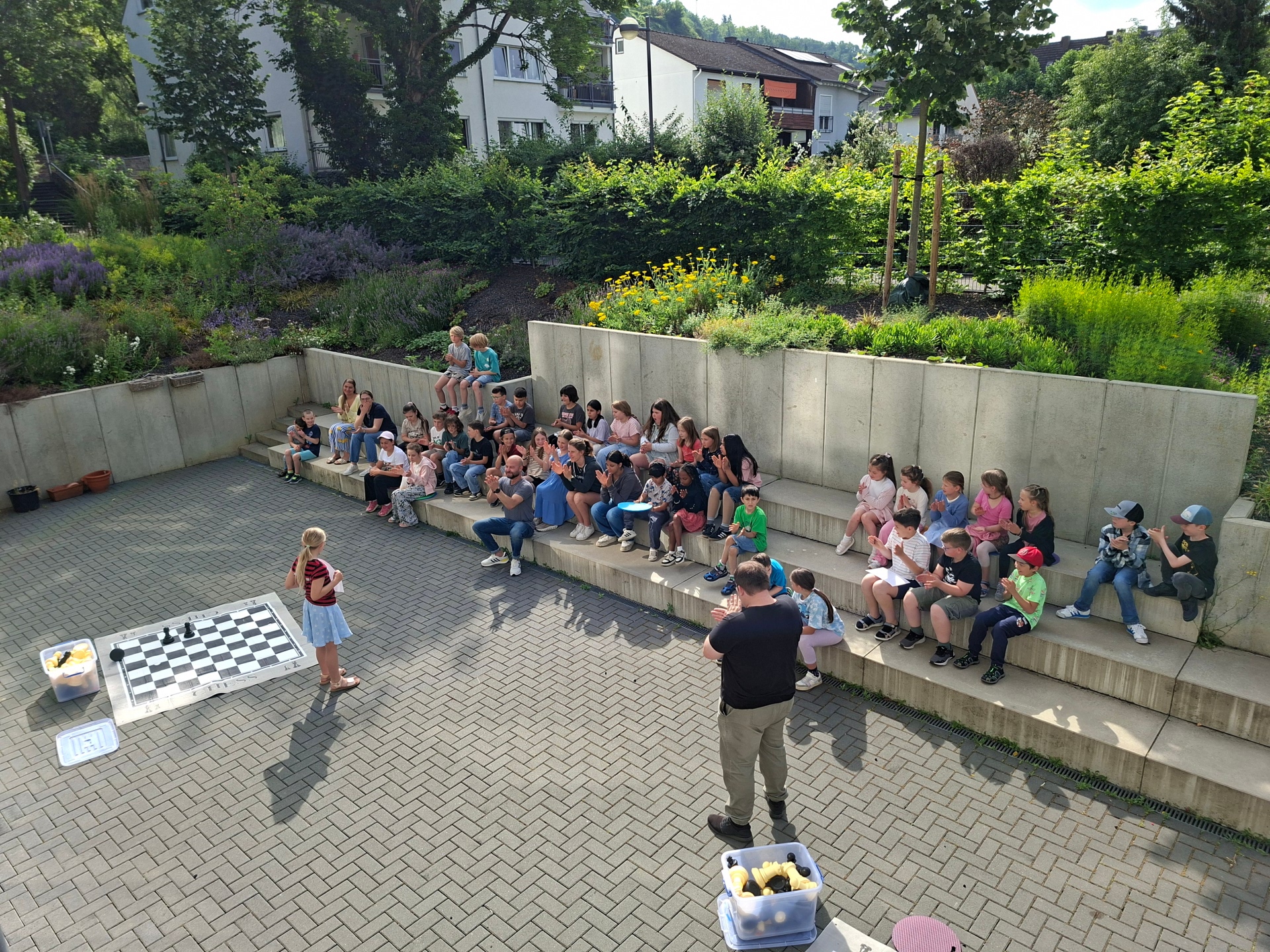 Eine Gruppe von Schülerinnen und Schülern sitzt im Freien auf einer breiten, terrassenförmigen Betonmauer. Im Vordergrund steht ein junges Mädchen vor einem großen Schachbrett auf dem Boden und hält ein Mikrofon, während ein Mann mit dem Rücken zur Kamera vor ihr steht. Die Kinder und Jugendlichen schauen aufmerksam zu und applaudieren teilweise, während sie in einer entspannten Umgebung mit viel Grün im Hintergrund zusammensitzen.