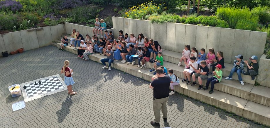 Goethe-Schule Lahnstein startet Talentprojekt für Schüler Eine Gruppe von Schülerinnen und Schülern sitzt im Freien auf einer breiten, terrassenförmigen Betonmauer. Im Vordergrund steht ein junges Mädchen vor einem großen Schachbrett auf dem Boden und hält ein Mikrofon, während ein Mann mit dem Rücken zur Kamera vor ihr steht. Die Kinder und Jugendlichen schauen aufmerksam zu und applaudieren teilweise, während sie in einer entspannten Umgebung mit viel Grün im Hintergrund zusammensitzen.