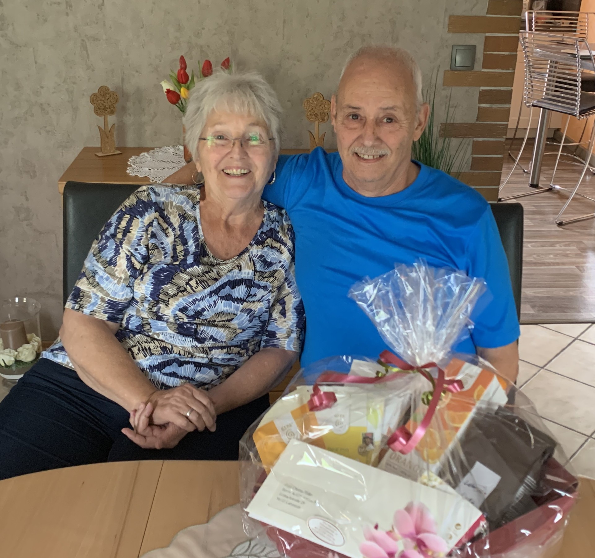 Ein älteres Ehepaar sitzt eng beieinander an einem Tisch in einem gemütlich eingerichteten Wohnraum und lächelt in die Kamera. Der Mann legt den Arm um die Frau, beide wirken glücklich und entspannt. Vor ihnen steht ein liebevoll verpackter Geschenkkorb mit Schleife und Karte, offenbar ein Präsent zu einem besonderen Anlass. Im Hintergrund sind eine Vase mit roten Tulpen sowie dekorative Elemente zu sehen, die eine warme, familiäre Atmosphäre schaffen.
