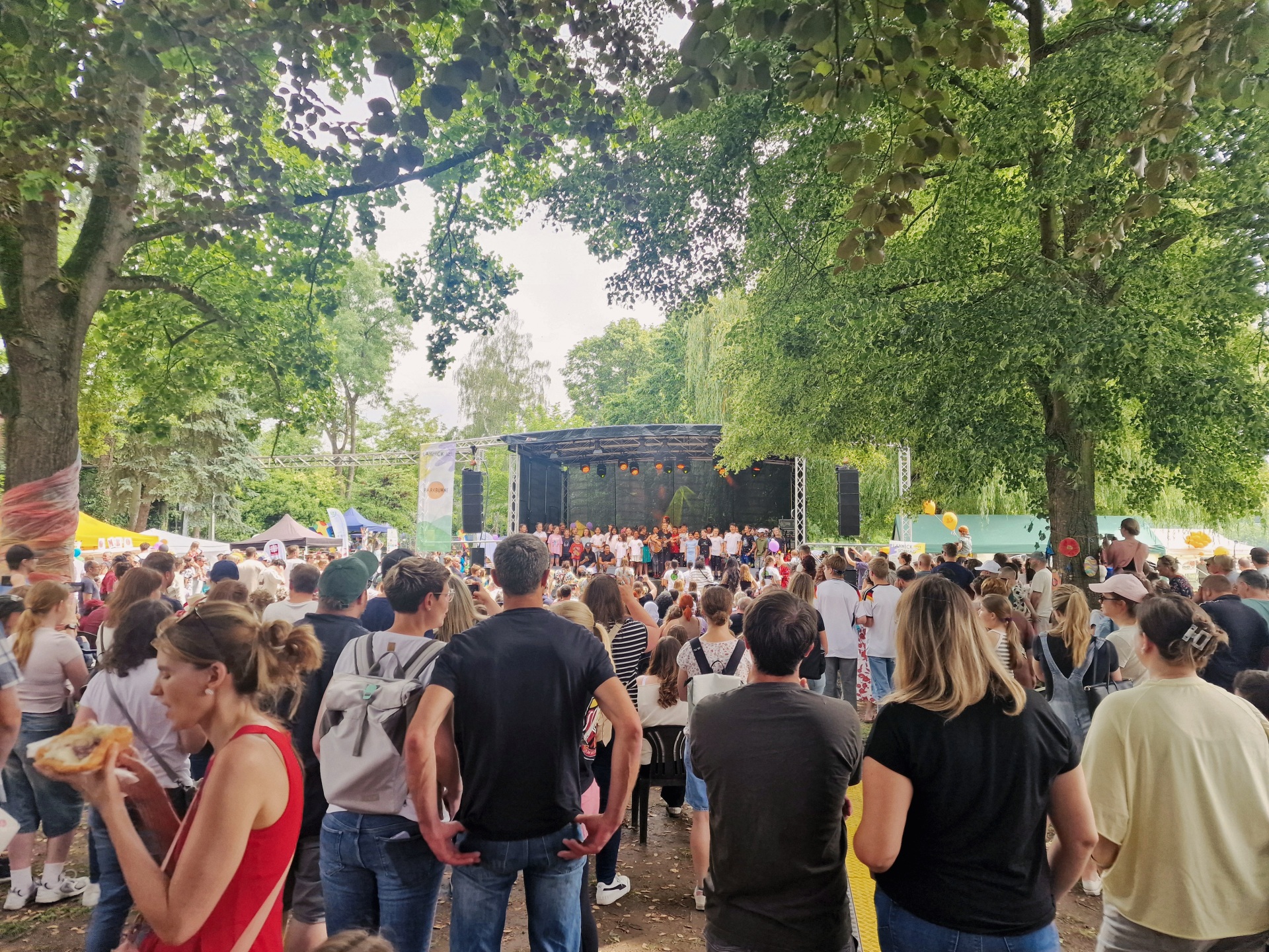 Zahlreiche Besucherinnen und Besucher verfolgen das Bühnenprogramm des Kinderprogramms von Lahneck Live in den Rheinanlagen Oberlahnstein. Vor der Bühne stehen und sitzen Familien unter großen schattenspendenden Bäumen, während Kinder und Jugendliche auf der Bühne auftreten.
