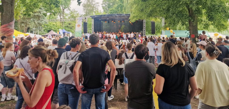 Zahlreiche Besucherinnen und Besucher verfolgen das Bühnenprogramm des Kinderprogramms von Lahneck Live in den Rheinanlagen Oberlahnstein. Vor der Bühne stehen und sitzen Familien unter großen schattenspendenden Bäumen, während Kinder und Jugendliche auf der Bühne auftreten.